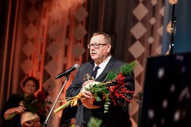 Vīrietis ar ziediem un balvu rokās runā mikrofona