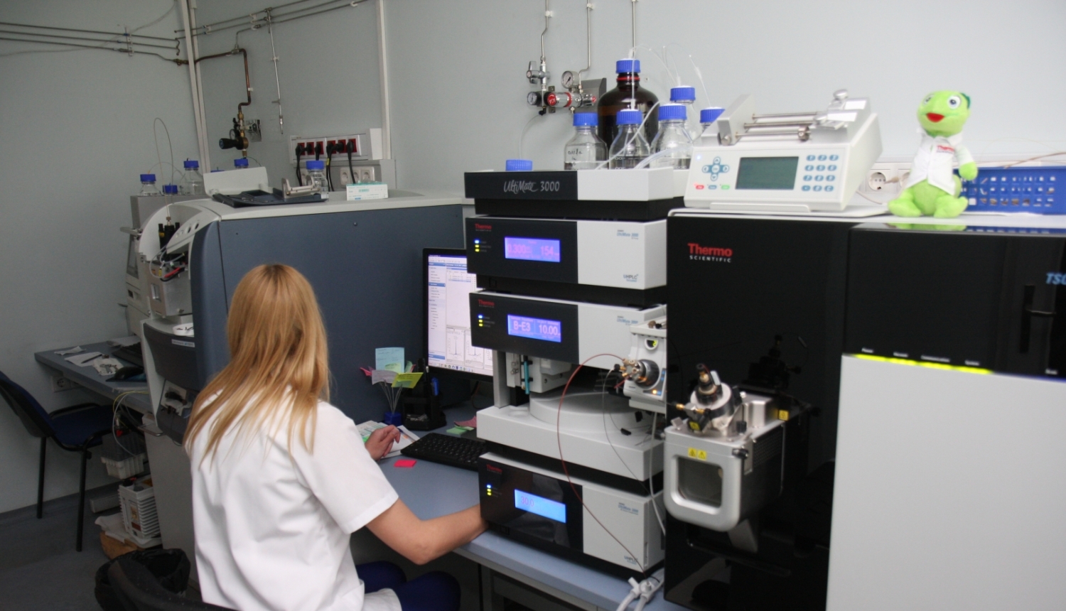Laboratorijas darbinieks sēž pie datora un strādā ar analītiskām iekārtām. 