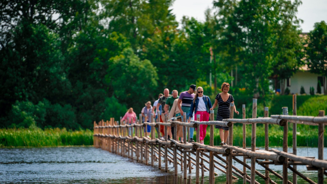 Pāri upei pa vienkāršu koka tiltu iet cilvēki zosu gājienā 