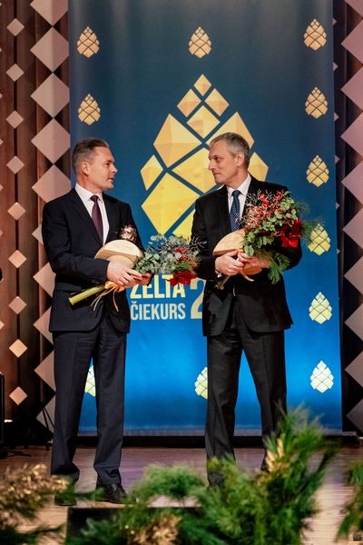 Divi vīrieši uz skatuves ar balvām un ziediem rokās