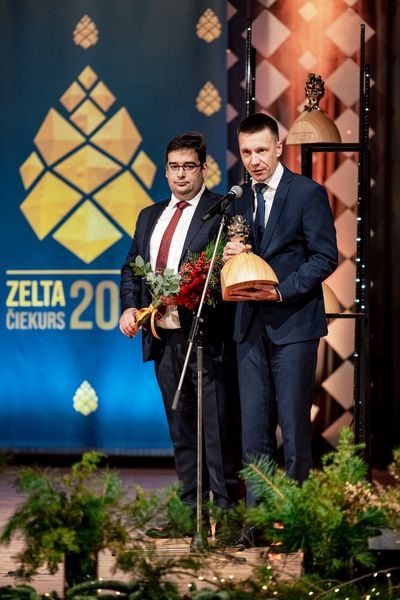 Divi vīrieši uz skatuves ar balvām un ziediem rokās, viens runā mikrofonā
