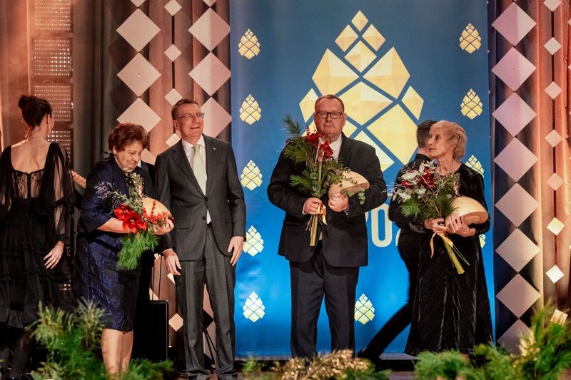 Uz skatuves Valsts prezidents ar sievietēm un vīriešim, kas tur rokās ziedus un balvas
