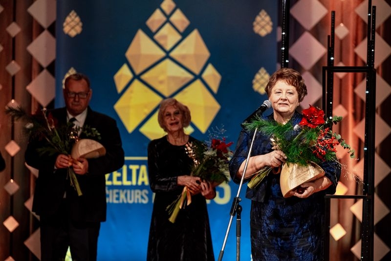 Pie mikrofona stāv sieviete ar balvu un ziediem rokās, fonā citi cilvēki ar balvām uz ziediem rokās
