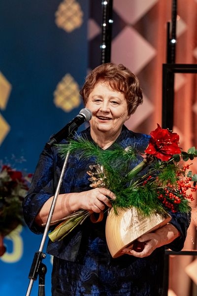 Sieviete runā pie mikrofona ar ziediem un balvu rokās