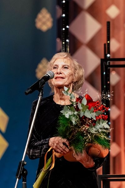 Sieviete runā pie mikrofona ar ziediem un balvu rokās
