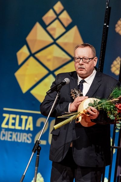 Vīrietis ar ziediem un balvu rokās runā mikrofona