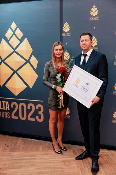 Pie zilas ar zelta čiekuriem foto sienas vīrietis, kas tur rokās diplomu un sieviete ar sarkaniem ziediem rokās