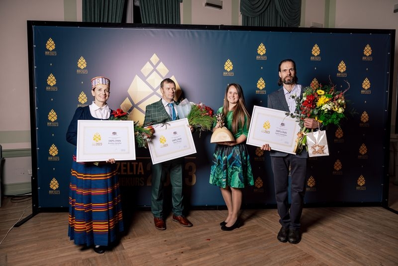 Pie zilas ar zelta čiekuriem foto sienas divi vīrieši un divas sievietes - visiem rokās ziedi un balvas