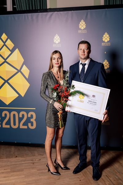 Pie zilas ar zelta čiekuriem foto sienas vīrietis, kas tur rokās diplomu un sieviete tur rokās sarkanus ziedus